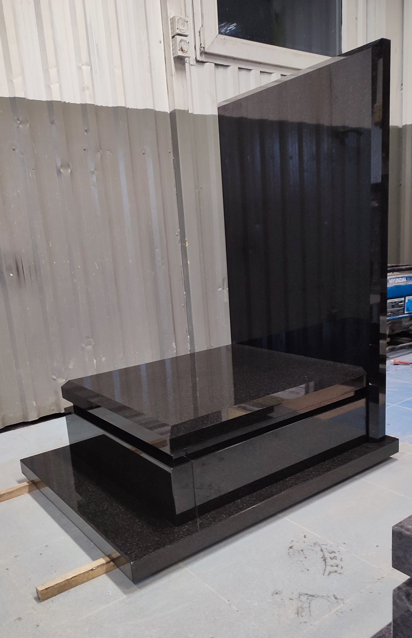 Surowy, czarny, granitowy pomnik z trzema poziomami, ustawiony w warsztacie na drewnianych legarach, z blachą falistą w tle.