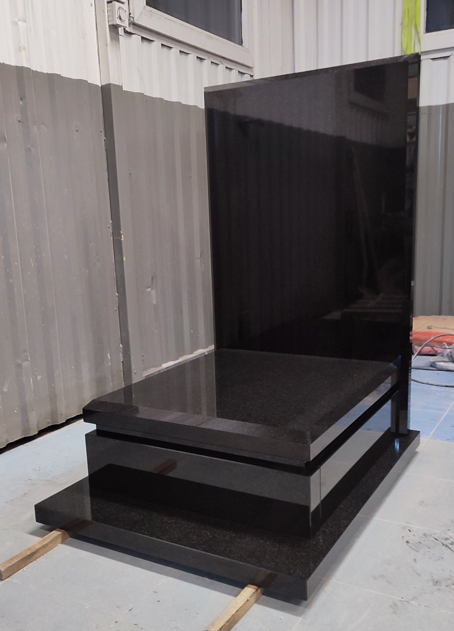 Nowoczesny, czarny, granitowy pomnik nagrobny z trzema poziomami, ustawiony w hali produkcyjnej na drewnianych podkładkach, przed ścianą z blachy falistej.