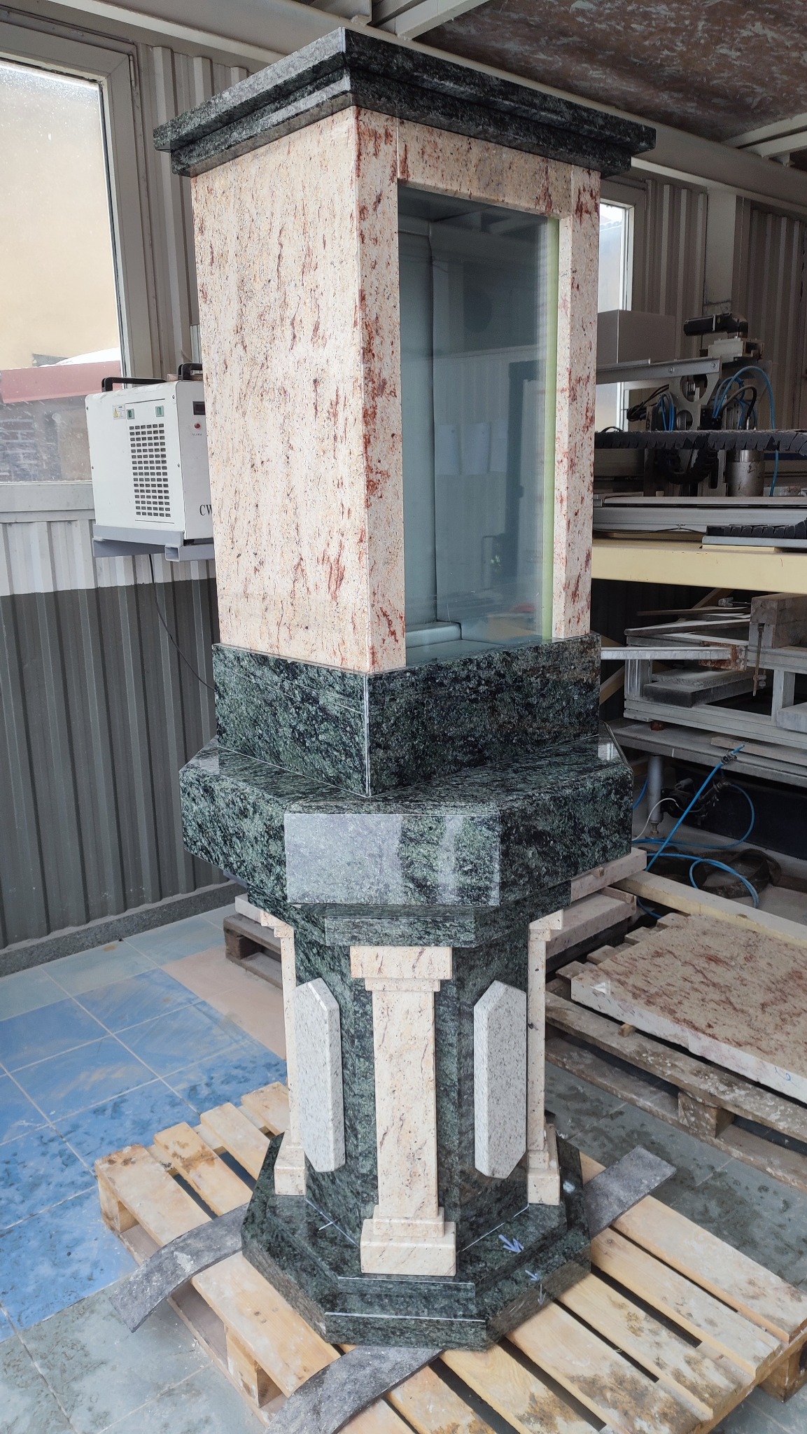 Wystawka z kamienia, z przeszkloną gablotą, wykonana w Strzegomiu, stojąca na palecie w warsztacie kamieniarskim. Elementy z zielonego i beżowego granitu.