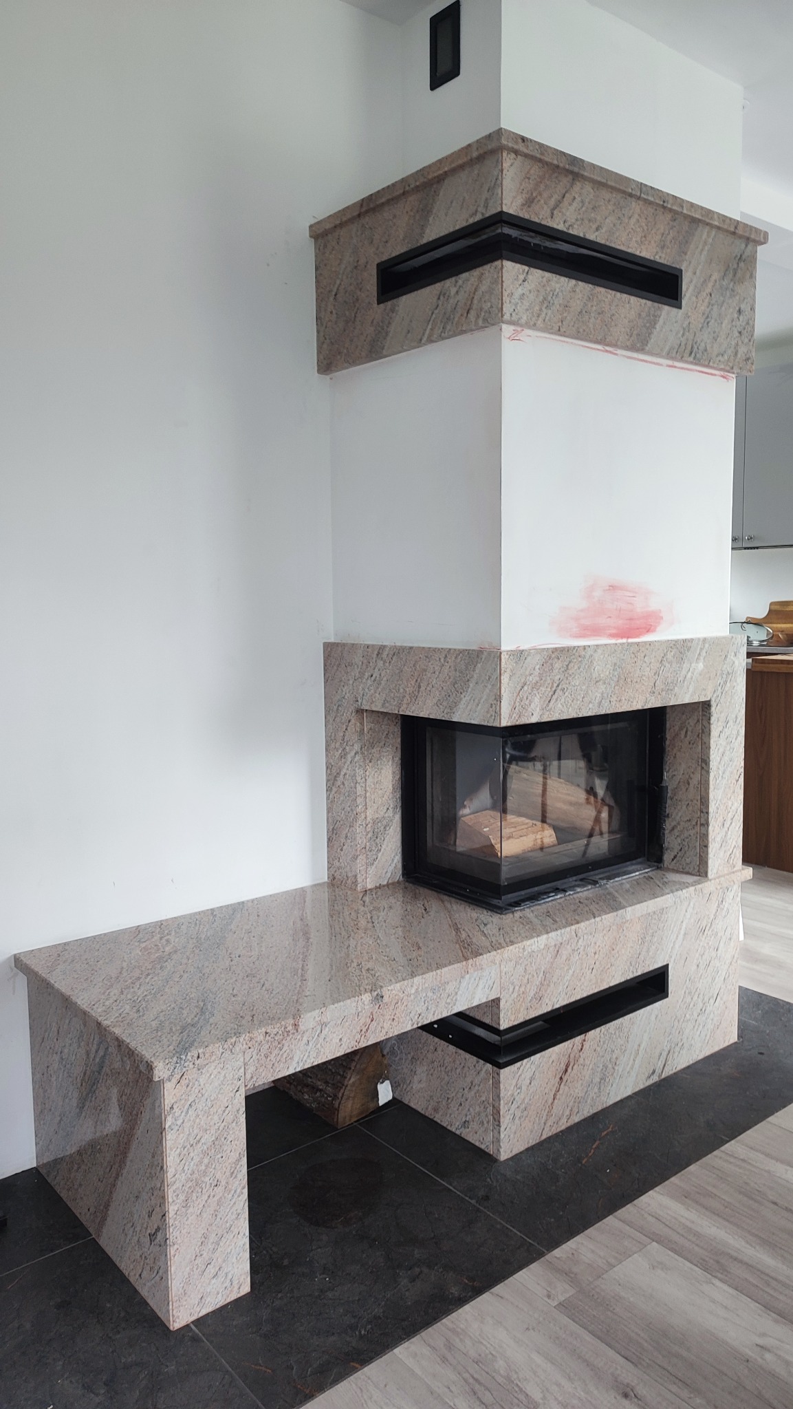 Kamienny kominek z blatem w odcieniach beżu i brązu, z czarnymi wstawkami. Widoczny wkład kominkowy i fragment podłogi z ciemnych płytek.