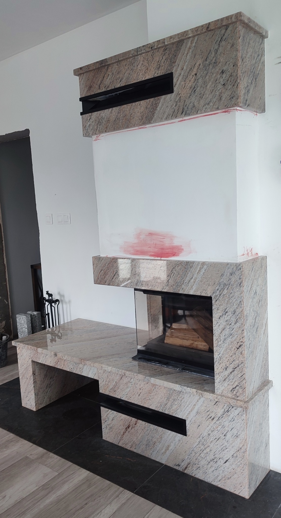 Kamienny kominek z przeszklonym paleniskiem i elementami z czarnego metalu. Widoczne ślady po malowaniu na ścianie. Całość w jasnej tonacji.