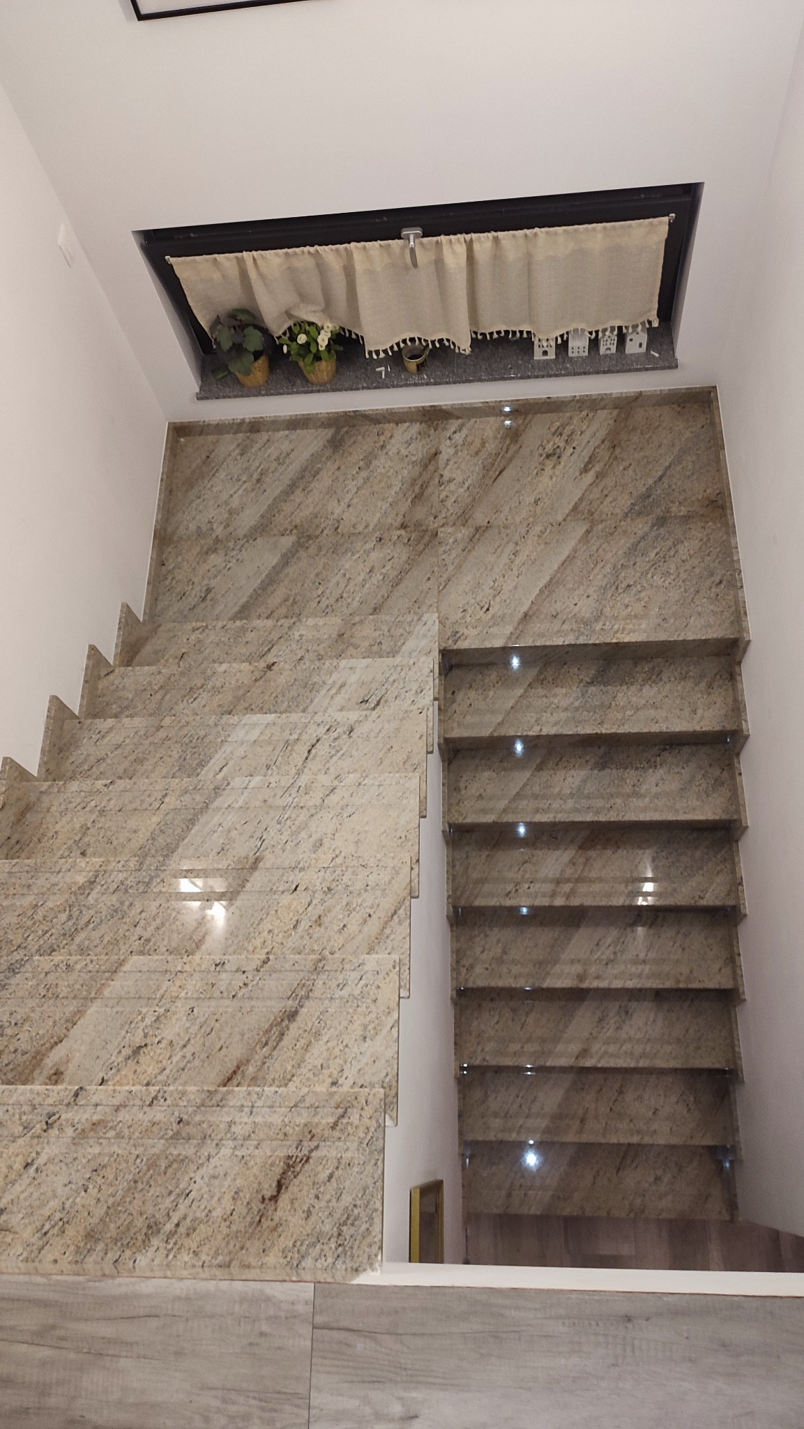 Granitowe schody wewnętrzne z podświetleniem LED, widok z góry. Dekoracyjny element nad schodami: rośliny i ozdobna zasłonka. Jasne, naturalne kolory kamienia.