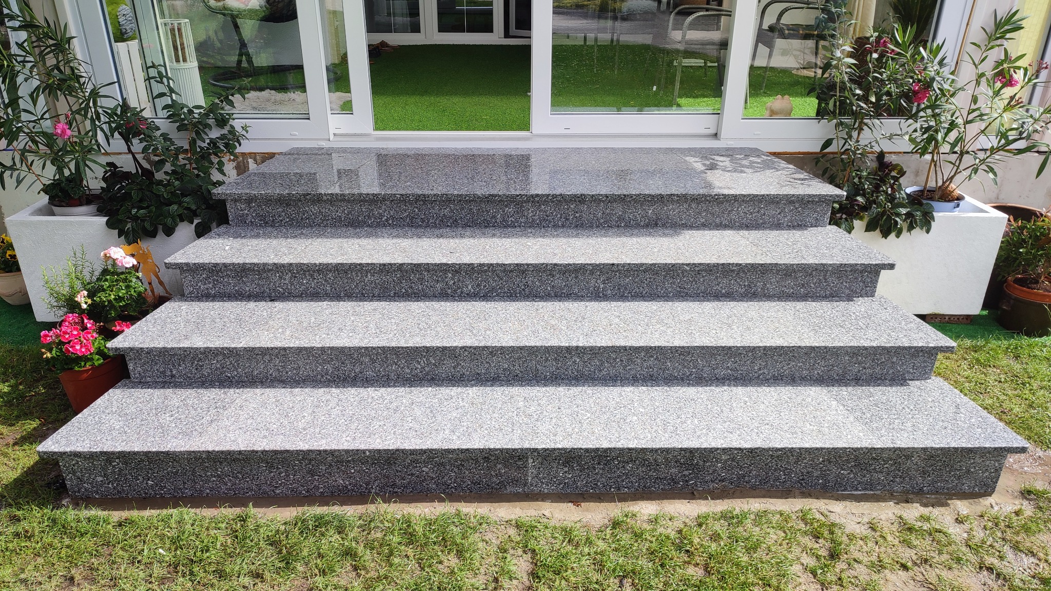Zewnętrzne schody z szarego kamienia prowadzące do domu, z roślinami w donicach po bokach. Widoczne cztery stopnie i fragment trawnika.