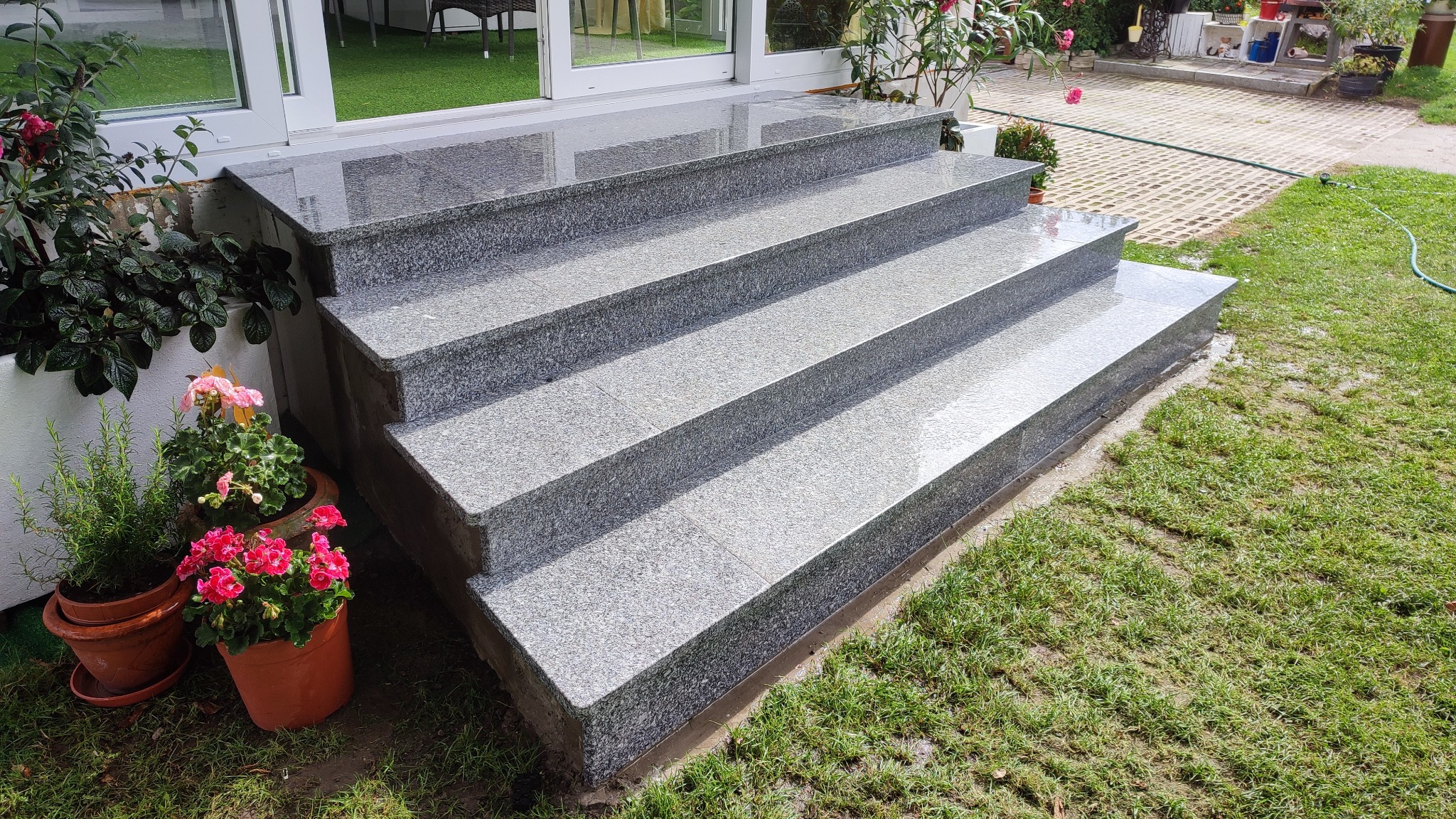 Granitowe schody zewnętrzne prowadzą do domu, otoczone zielenią trawnika i doniczkami z kwiatami. Polerowana powierzchnia schodów odbija światło.