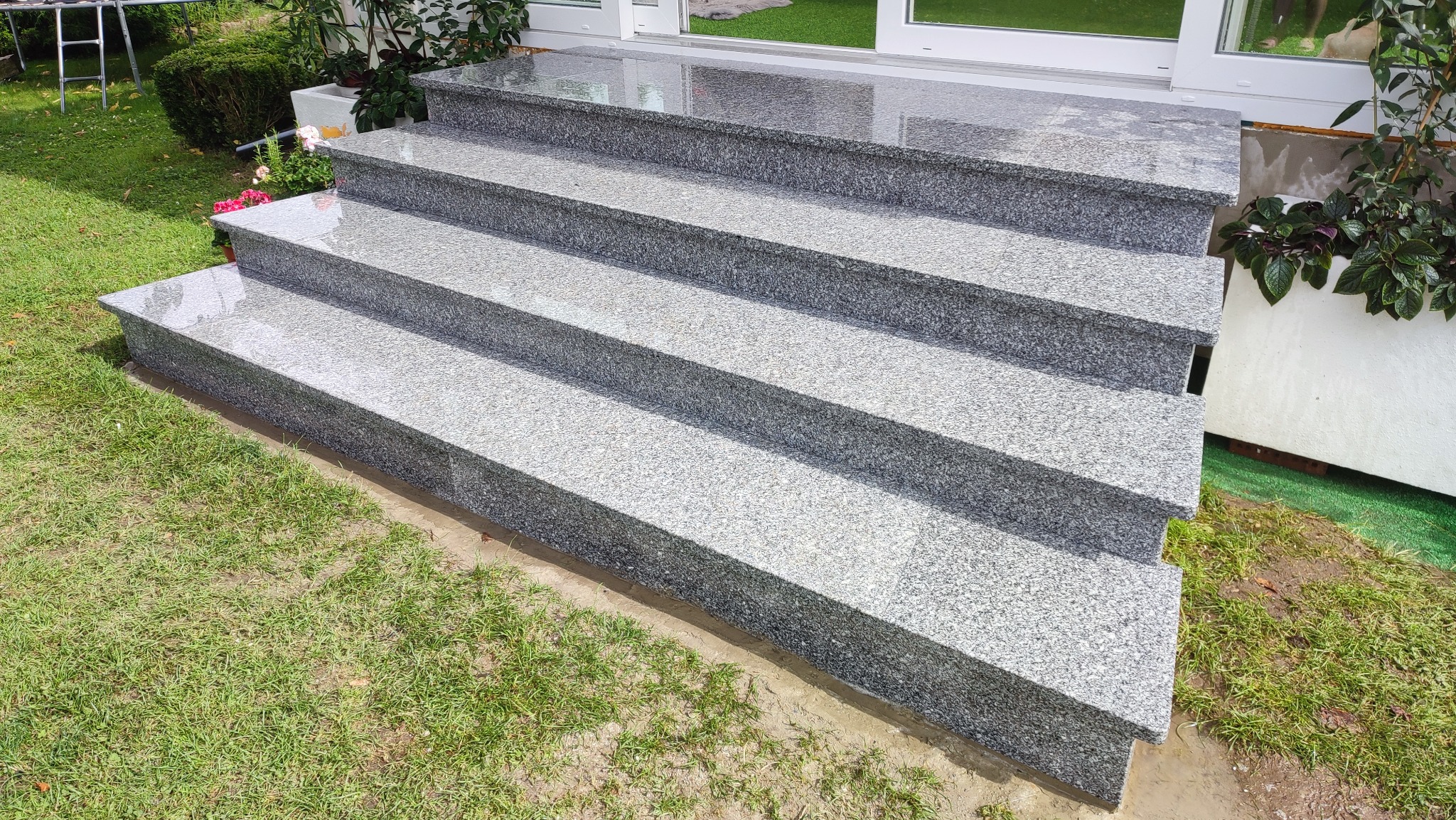 Zewnętrzne schody z szarego, ziarnistego kamienia prowadzące do drzwi tarasowych, otoczone zielenią trawnika i roślin w donicach. Widok z dołu.