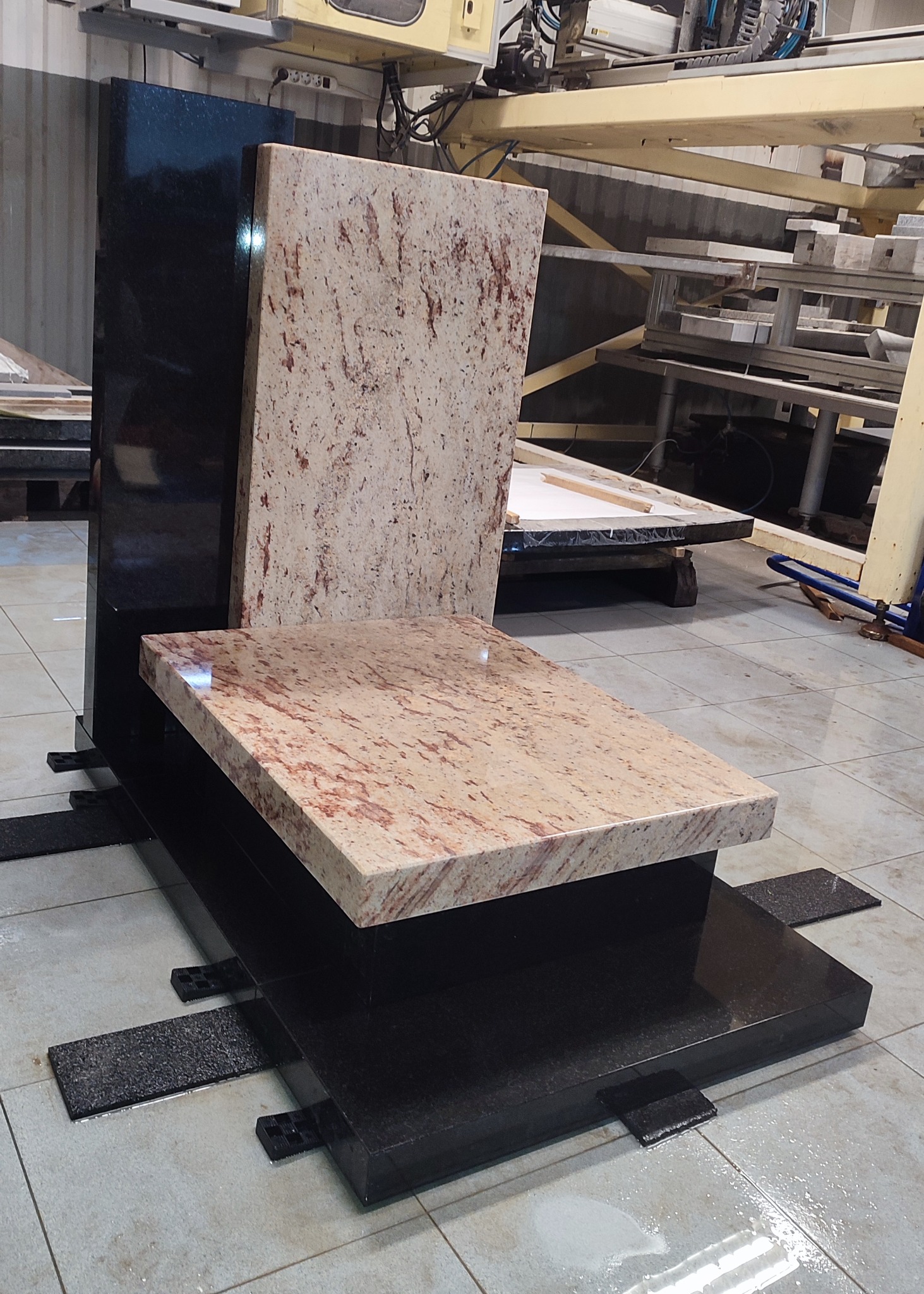 Nowoczesny nagrobek z jasnego i ciemnego kamienia, widoczny w warsztacie kamieniarskim, z elementami mocującymi na podłodze. Jasny blat z wyraźnym wzorem.