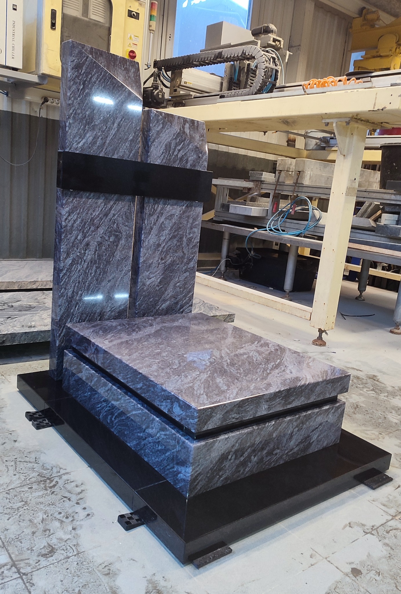 Granitowy nagrobek w odcieniach szarości i czerni, z widoczną strukturą kamienia, ustawiony w warsztacie kamieniarskim z maszyną do obróbki kamienia w tle.