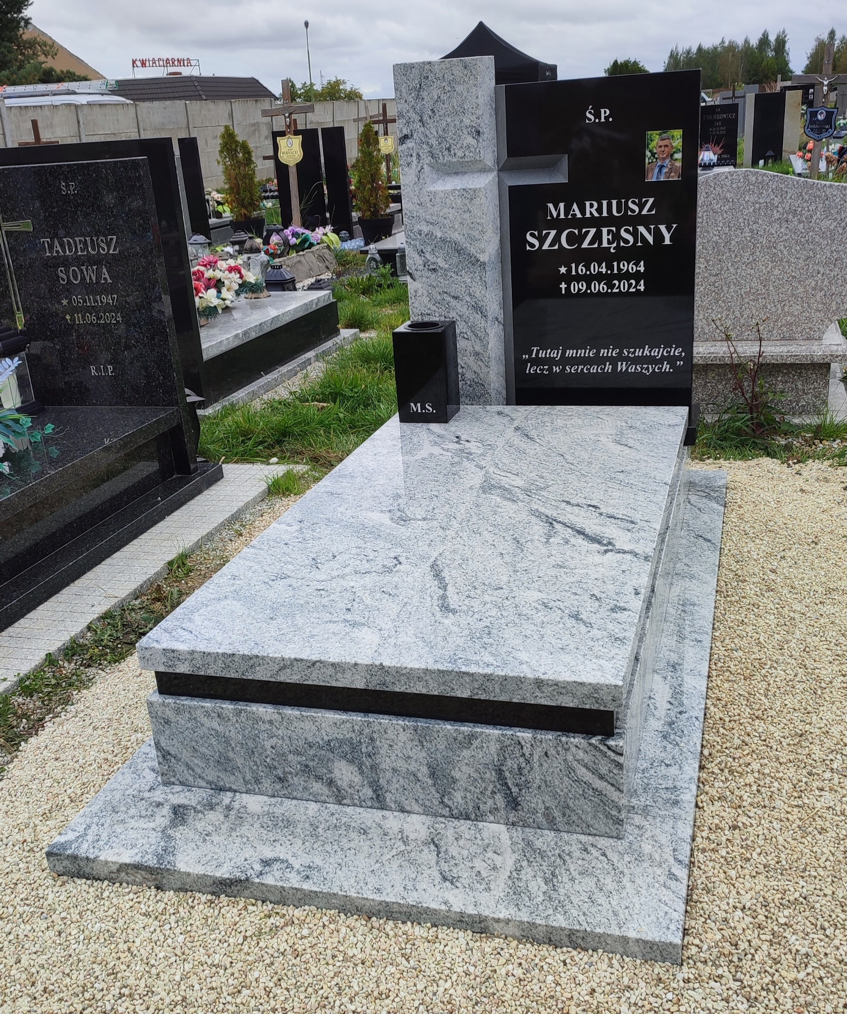 Granitowy nagrobek z czarną tablicą i zdjęciem osoby zmarłej, otoczony żwirem na cmentarzu. Widoczne inne nagrobki w tle, pochmurne niebo.