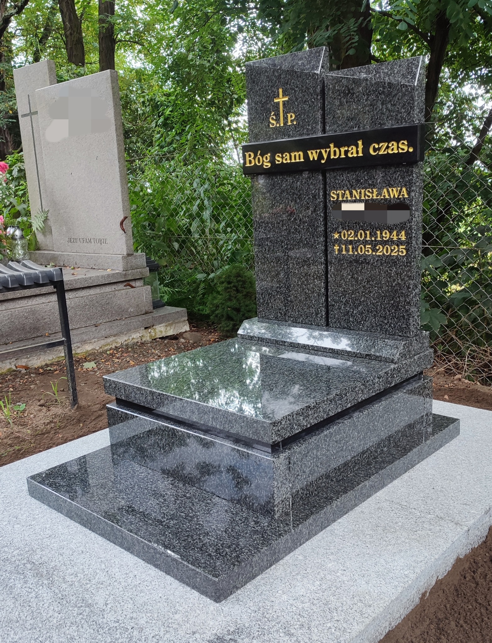 Granitowy nagrobek z wygrawerowanym napisem 'Bóg sam wybrał czas' i datami urodzin i śmierci, na tle zieleni cmentarza. Polerowana powierzchnia odbija otoczenie.