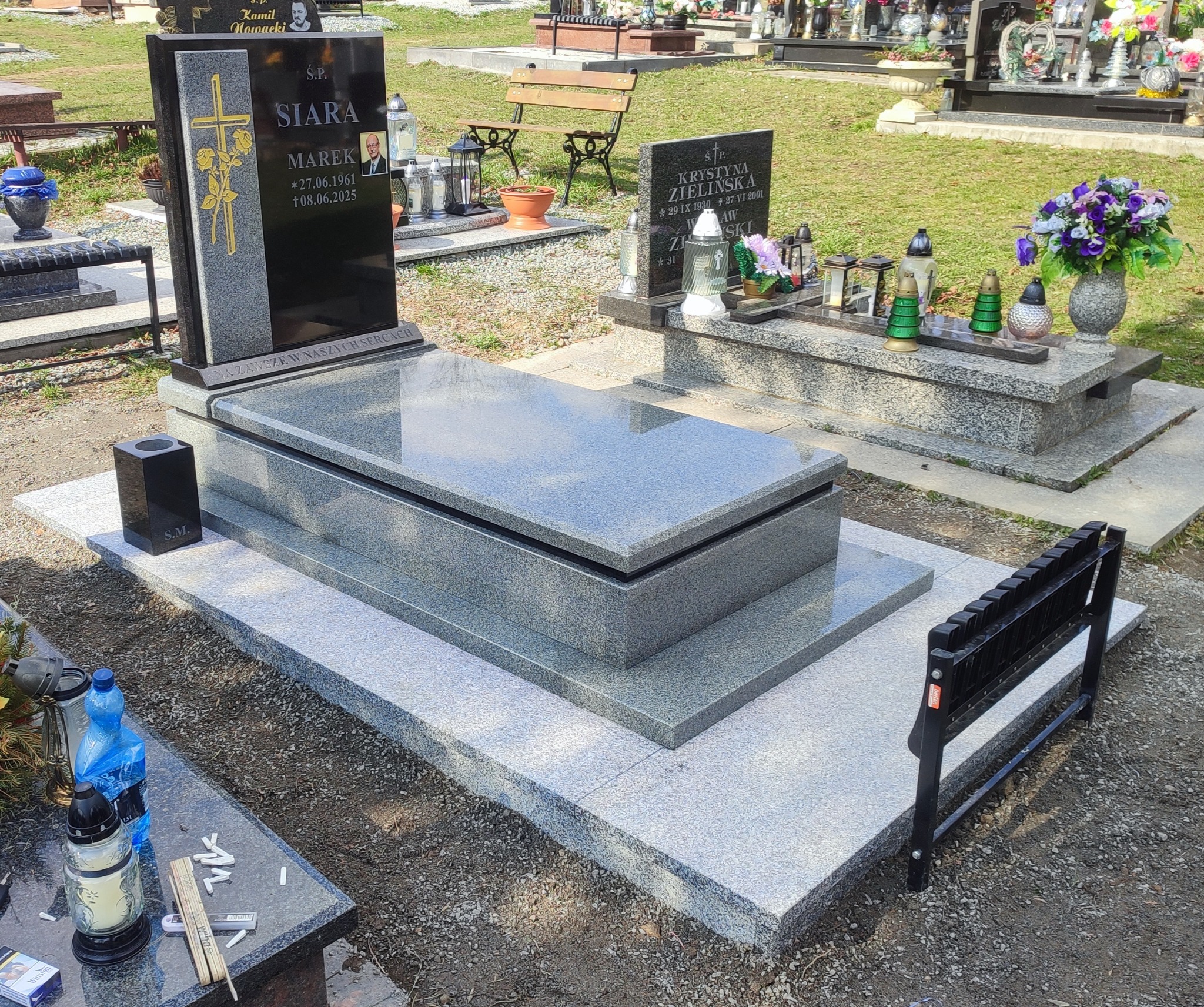 Granitowy nagrobek z czarną tablicą i złotym krzyżem na cmentarzu. Widoczny wazon na kwiaty i ławka. Nagrobek z inskrypcjami i datami.