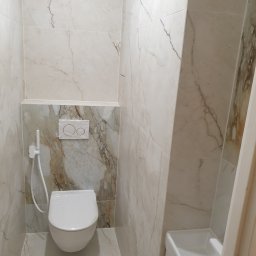 Kraszbud - Wnętrze małej łazienki z wiszącą toaletą owiniętą folią ochronną, bidetką i umywalką w rogu, wykończone płytkami imitującymi marmur z delikatnymi żyłkami.