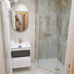 Kraszbud - Nowoczesna łazienka z kabiną prysznicową z elementami w kolorze złotym, umywalką z szafką i lustrem owalnym, w którym odbija się osoba robiąca zdjęcie.