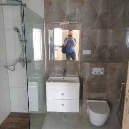 Kraszbud - Nowoczesna łazienka z kabiną prysznicową ze szklaną ścianką, umywalką z szafką, podwieszaną toaletą i marmurowymi płytkami na ścianach, sfotografowana z odbiciem osoby robiącej zdjęcie w lustrze.