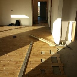 Kraszbud - Montaż drewnopodobnych płytek podłogowych w jasnym wnętrzu, widoczny poziomica, krzyżyki dystansowe i częściowo ułożona podłoga.