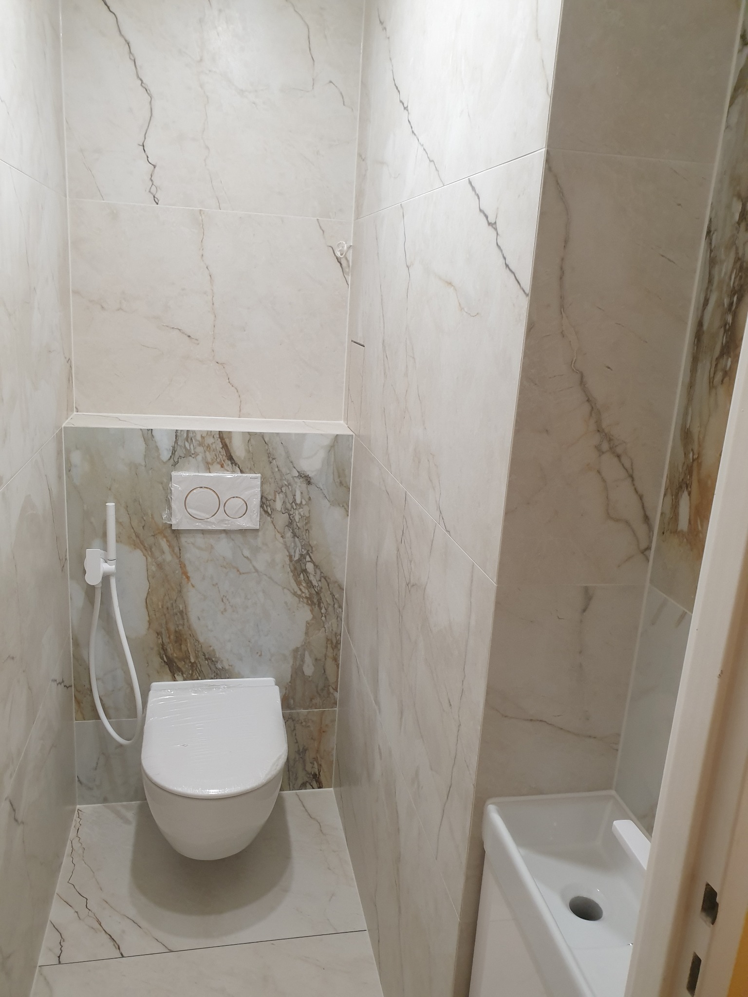 Wnętrze małej łazienki z wiszącą toaletą owiniętą folią ochronną, bidetką i umywalką w rogu, wykończone płytkami imitującymi marmur z delikatnymi żyłkami.