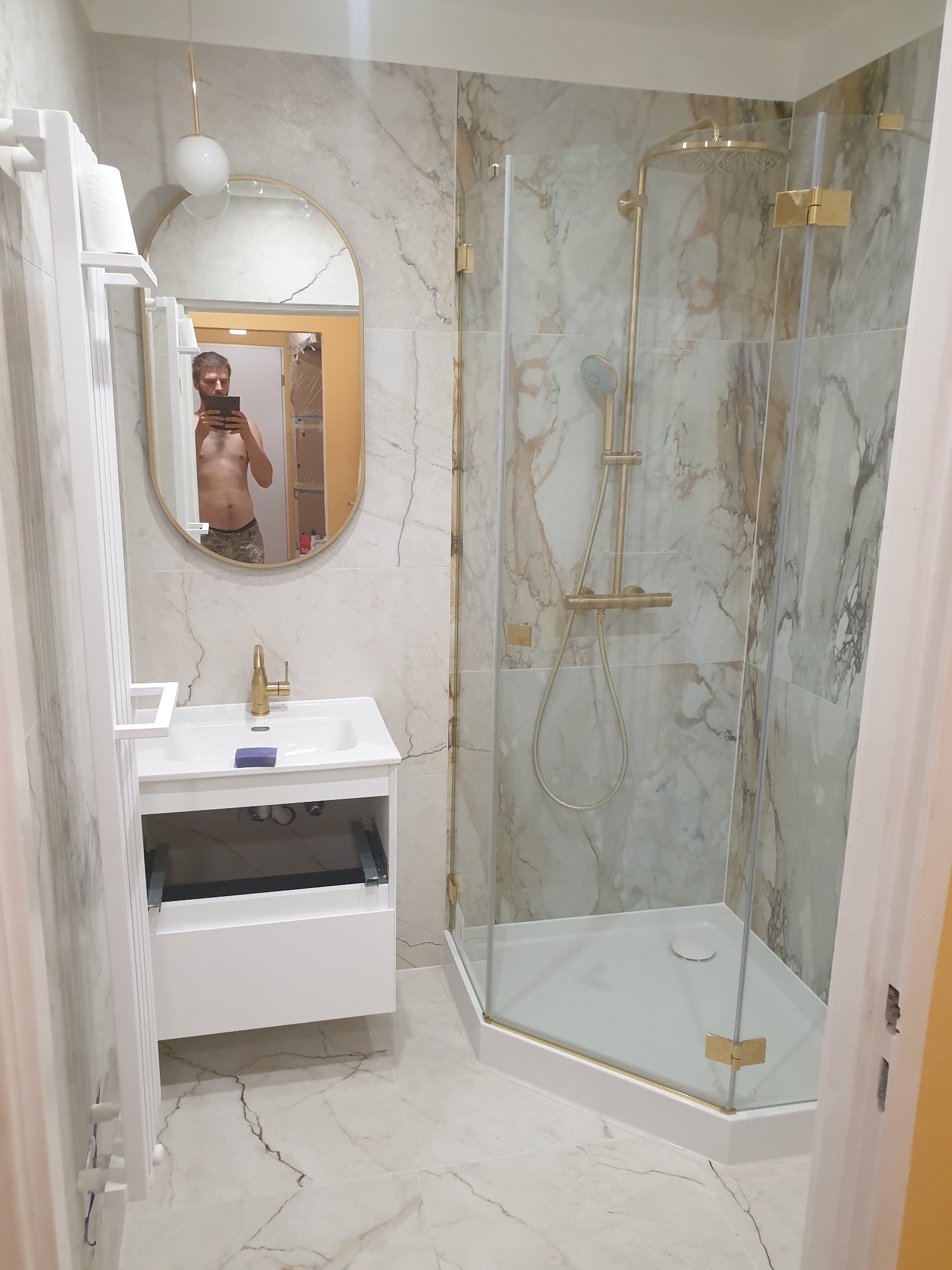 Nowoczesna łazienka z kabiną prysznicową z elementami w kolorze złotym, umywalką z szafką i lustrem owalnym, w którym odbija się osoba robiąca zdjęcie.