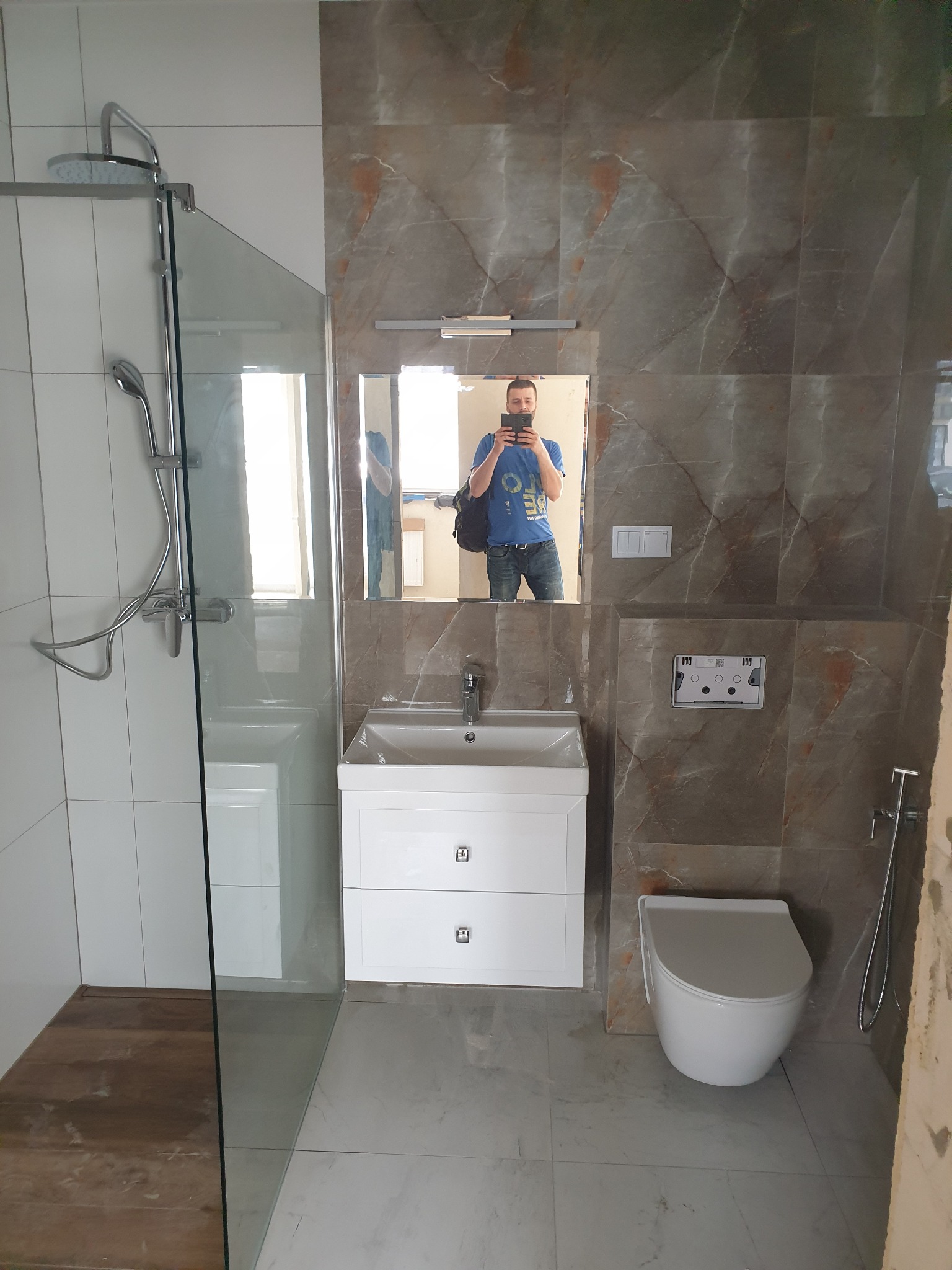 Nowoczesna łazienka z kabiną prysznicową ze szklaną ścianką, umywalką z szafką, podwieszaną toaletą i marmurowymi płytkami na ścianach, sfotografowana z odbiciem osoby robiącej zdjęcie w lustrze.