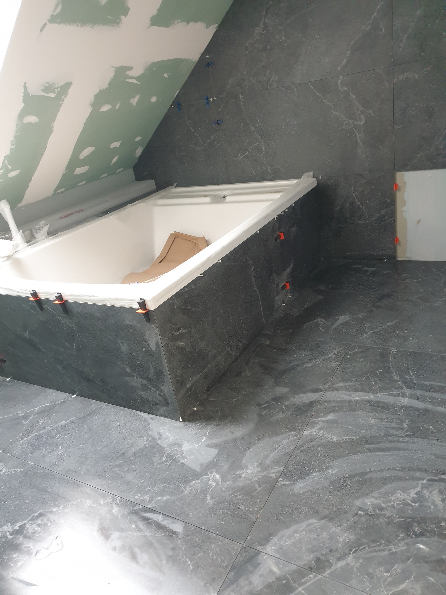 Montaż szarych płytek imitujących marmur wokół wanny w łazience na poddaszu, widoczne krzyżyki dystansowe i niezakończone szpachlowanie skosu.