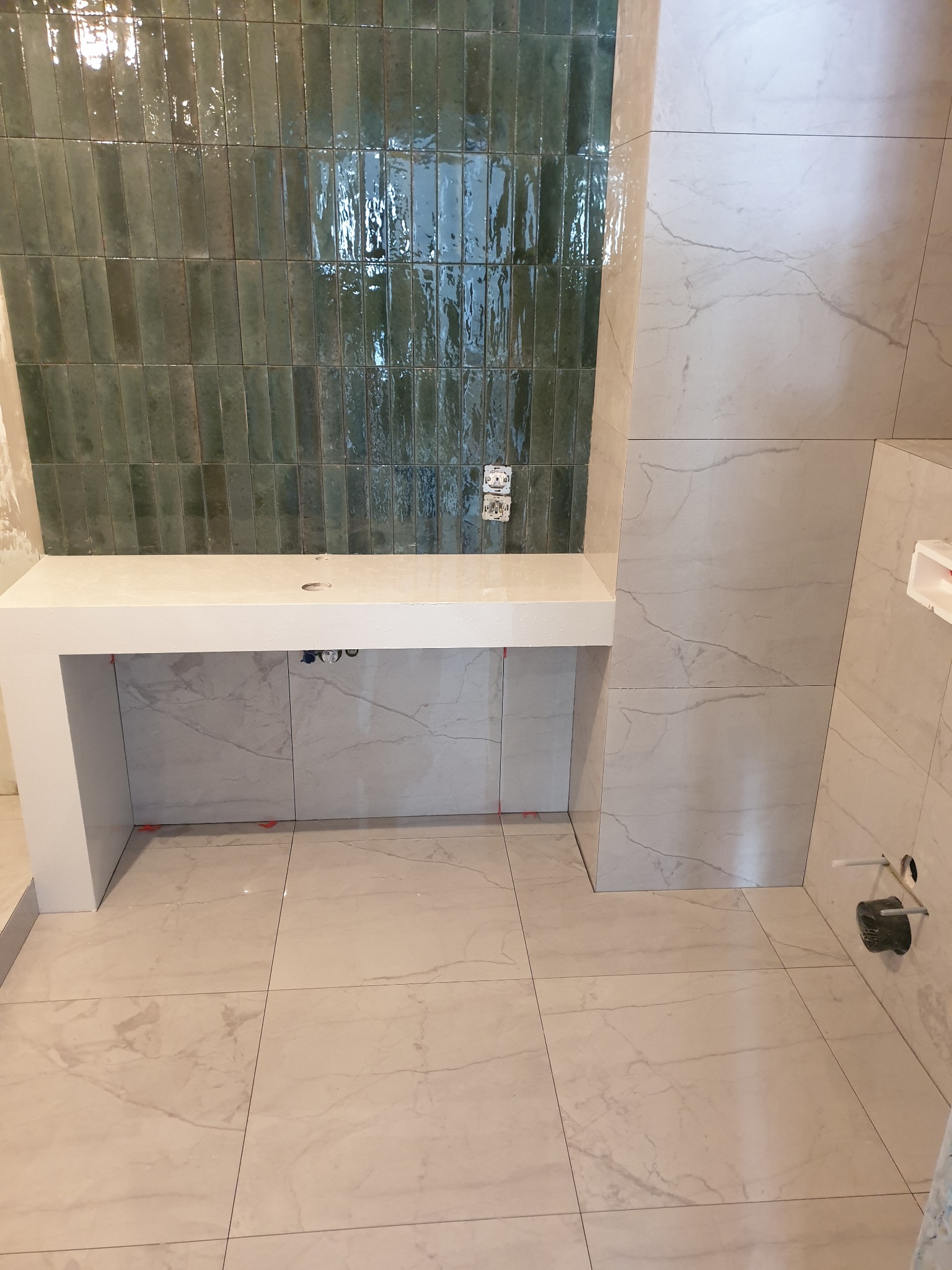 Świeżo położone, błyszczące, pionowe, zielone płytki na ścianie w łazience, kontrastujące z jasnymi, marmurkowymi płytkami na podłodze i ścianach oraz białą zabudową pod umywalkę z widocznymi...