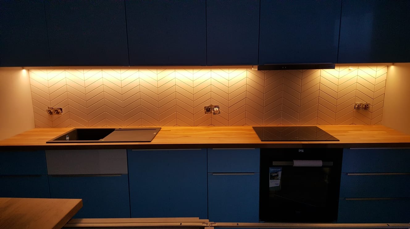 Nowoczesna kuchnia z niebieskimi szafkami, drewnianym blatem i biało-beżową mozaiką ułożoną w jodełkę nad blatem, podświetloną listwą LED.