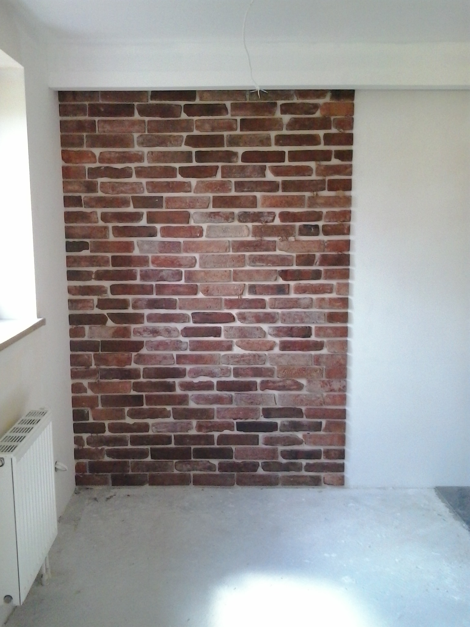 Częściowo wykończona ściana z cegły dekoracyjnej we Wrocławiu, sąsiadująca z gładką, pomalowaną na biało ścianą. Widoczny grzejnik i surowa podłoga.