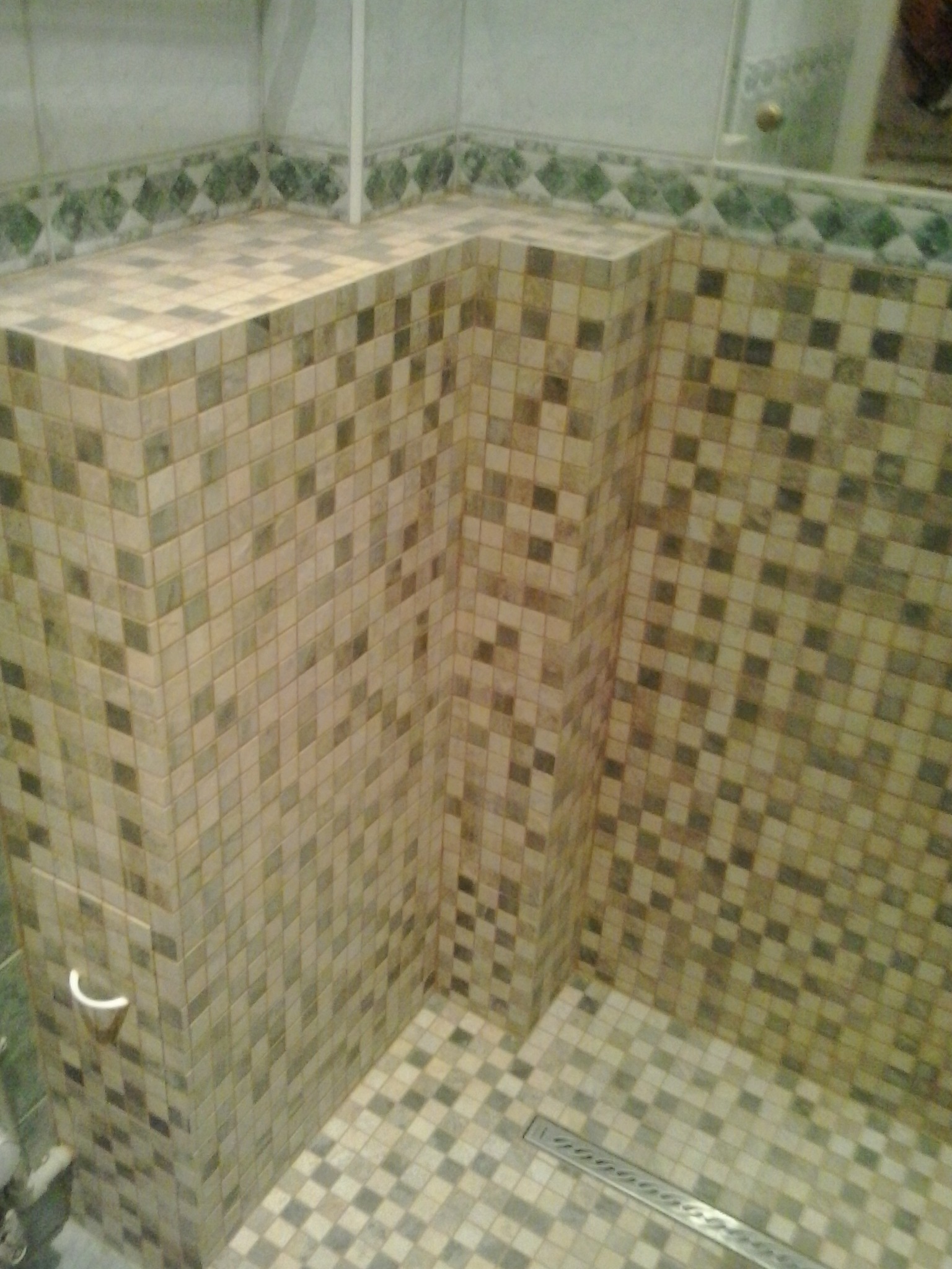 Wykończona kabina prysznicowa z mozaiką w odcieniach beżu i szarości, widoczny odpływ liniowy i uchwyt na ręcznik, dekoracyjny pas płytek na ścianie w tle.