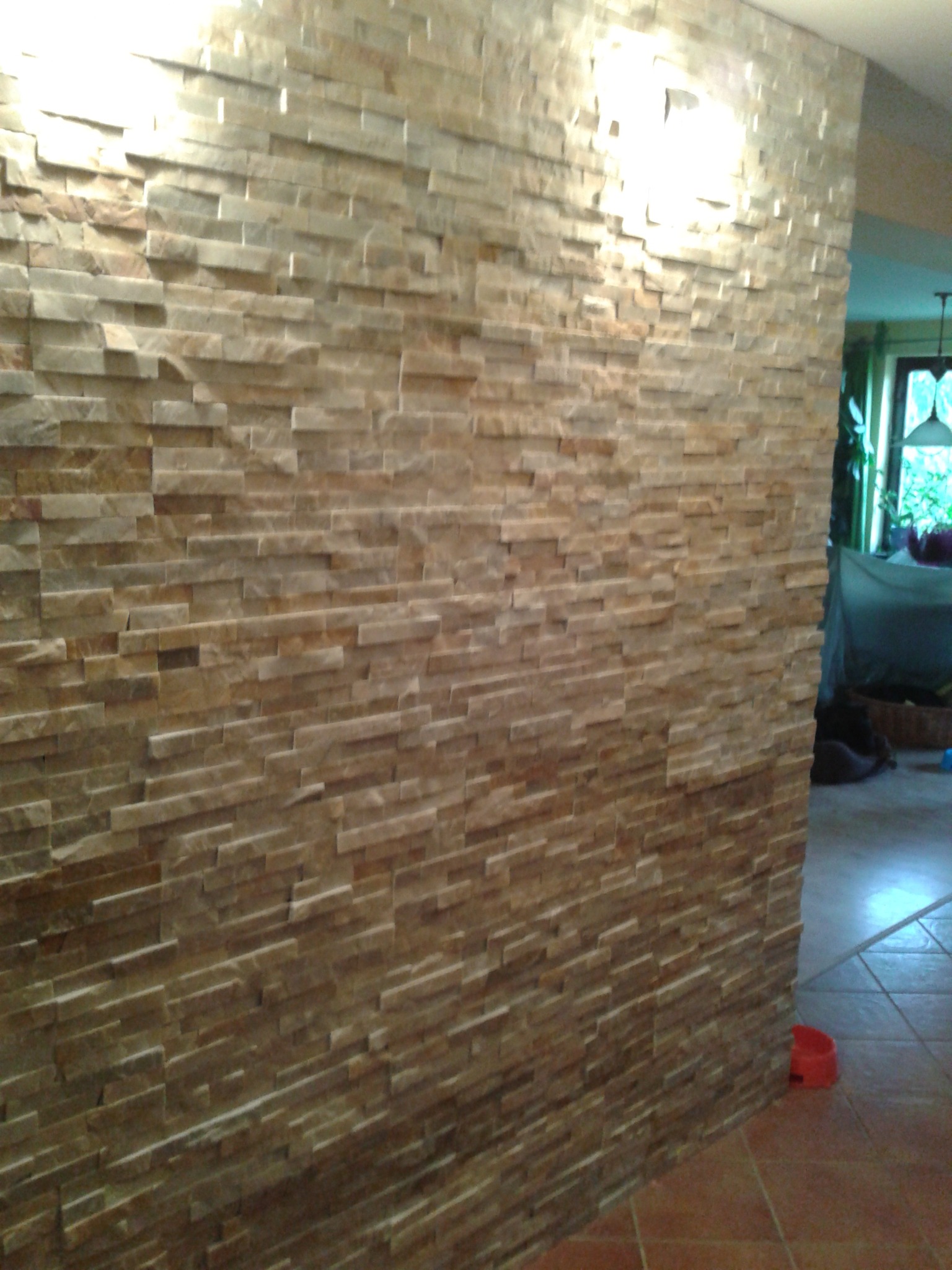 Ściana wykończona kamieniem dekoracyjnym w odcieniach beżu i brązu, z widocznym oświetleniem punktowym i fragmentem wnętrza z terakotą na podłodze oraz czerwoną miską dla zwierząt.