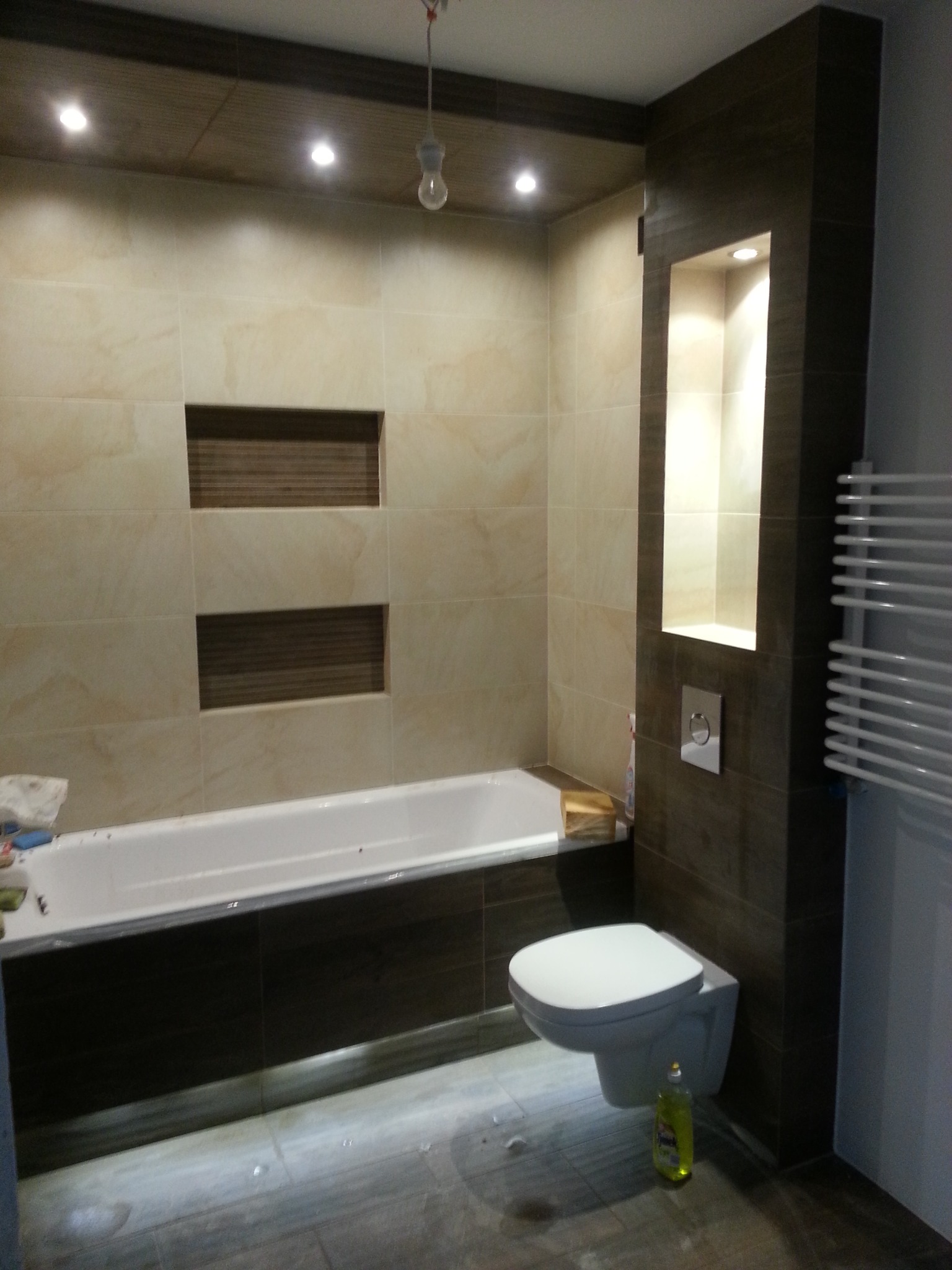 Wykończona łazienka z beżowymi płytkami, wnękami w ścianie nad wanną i podświetlaną wnęką obok toalety. Na podłodze widoczne ślady po remoncie, a na wannie leżą narzędzia.