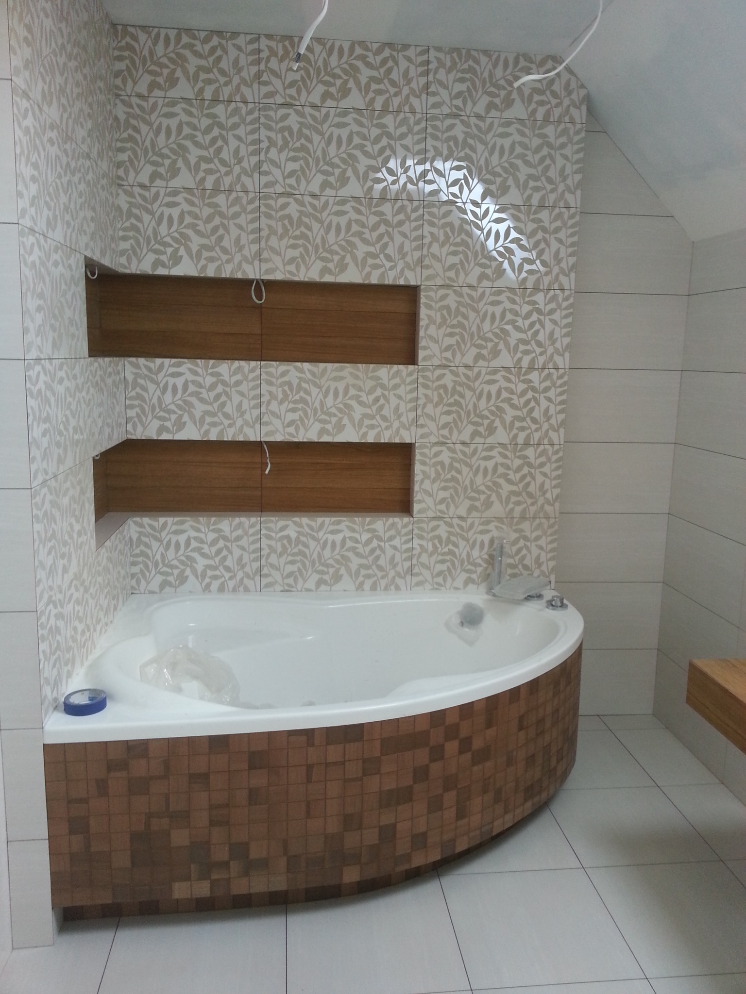 Nowoczesna łazienka z wanną obudowaną mozaiką w odcieniach brązu, ściany wyłożone płytkami z motywem liści, w ścianie wnęki wykończone drewnem, widoczne niedokończone podłączenie elektryczne.