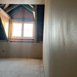 Omitky Rafał Kabut - Poddasze po nałożeniu gładzi gipsowej, widoczne drewniane elementy konstrukcji dachu i okno dachowe z wpadającym światłem słonecznym.