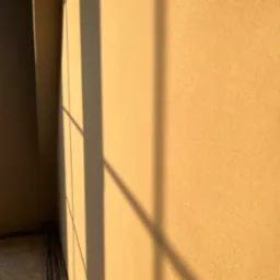 Ściana pokryta świeżym, jasnym tynkiem, oświetlona popołudniowym słońcem, rzucającym cienie okiennej ramy.