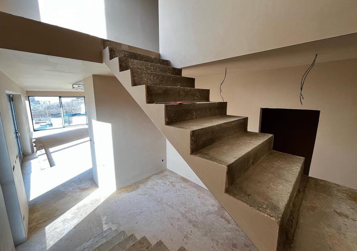 Surowe schody betonowe w nowo otynkowanym wnętrzu, widoczne ślady po pracach budowlanych, wystające przewody elektryczne i duże okno z widokiem na zewnątrz.