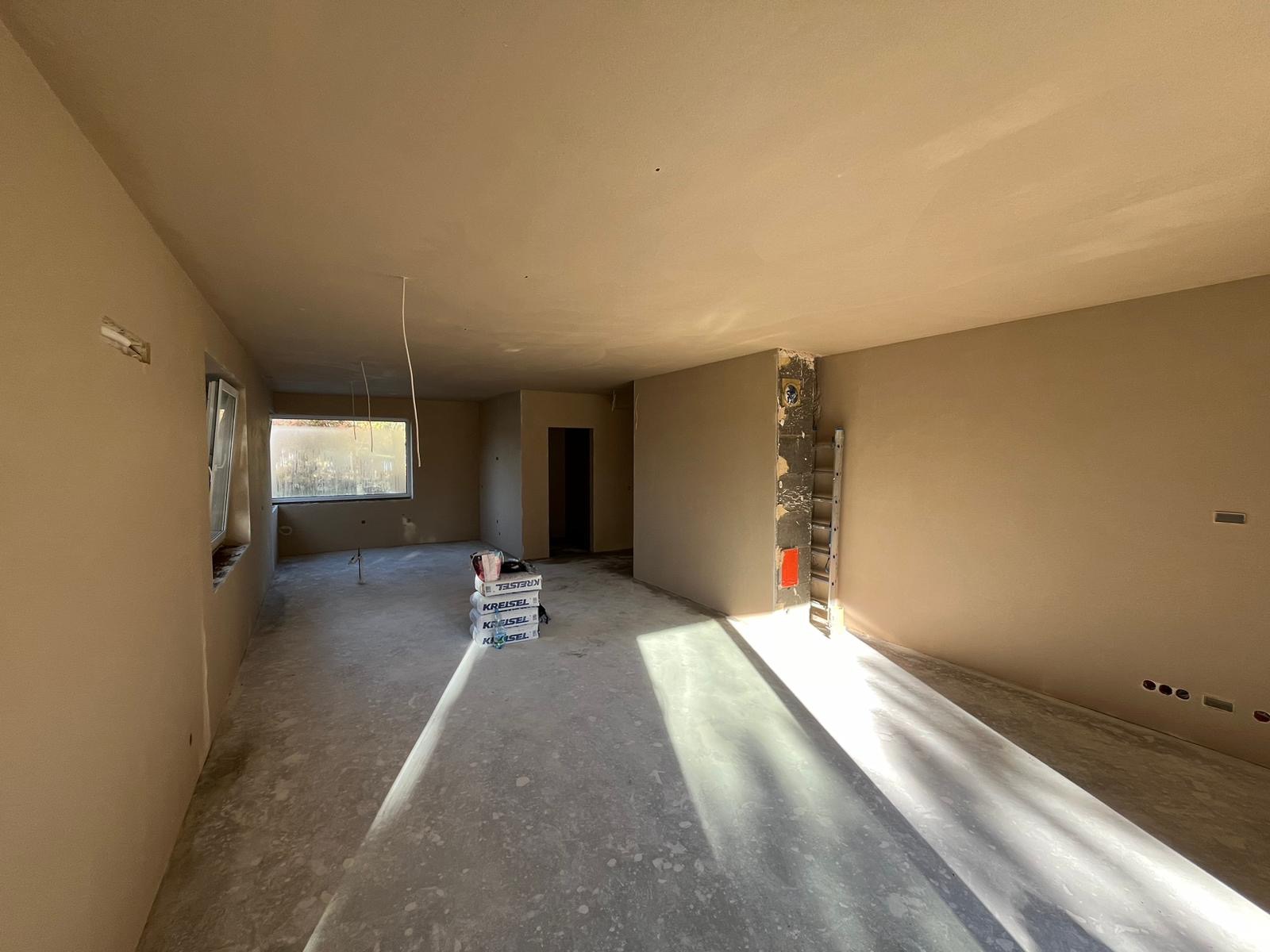 Wnętrze po tynkowaniu maszynowym, widoczne ściany i sufit pokryte świeżym tynkiem, na podłodze paleta z workami z zaprawą, w tle okno i drabina oparta o ścianę.