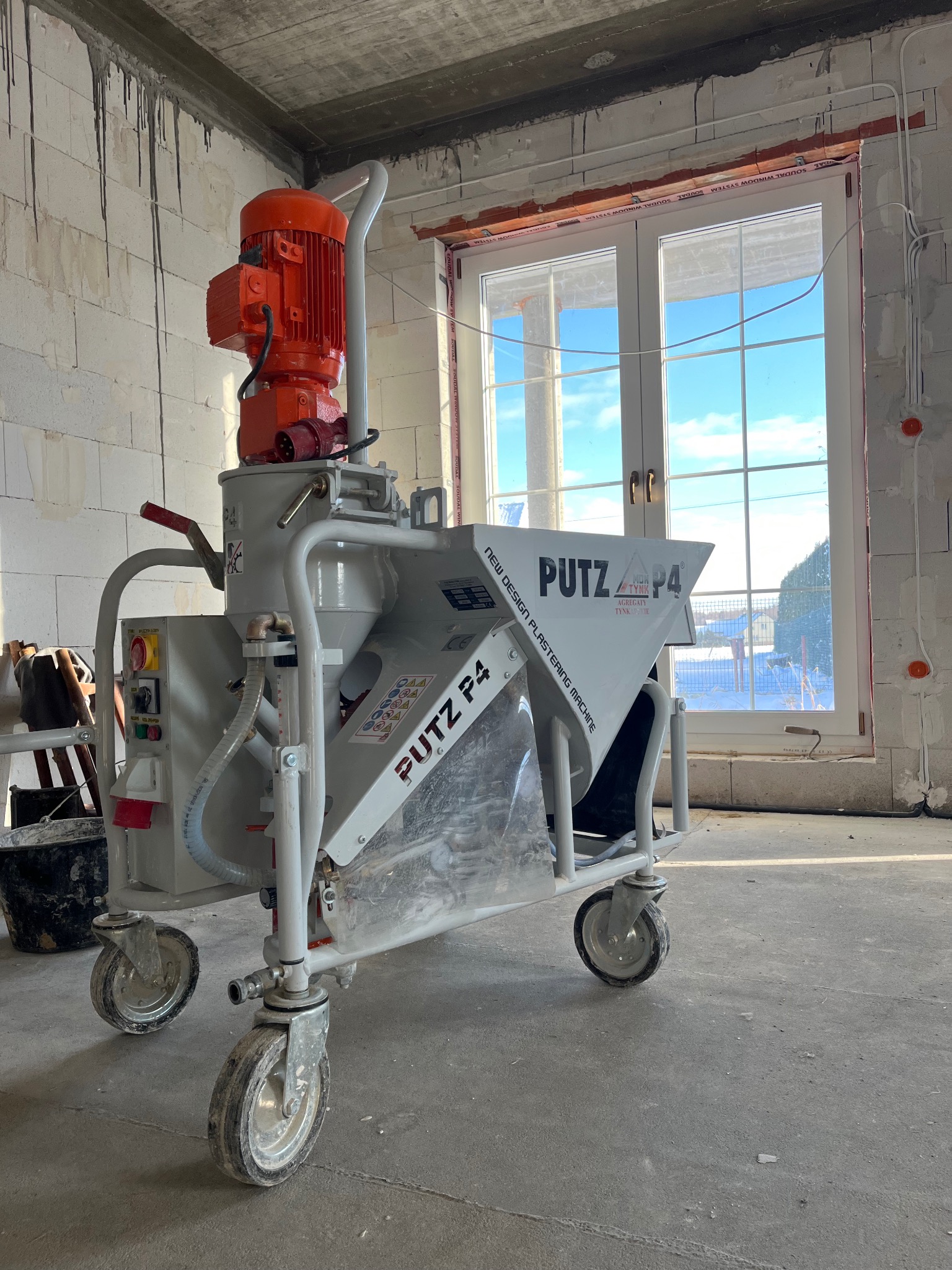 Agregat tynkarski P4 marki Putz w surowym wnętrzu, widoczny w oknie błękit nieba, ściany z pustaków z widocznymi śladami po zaprawie.