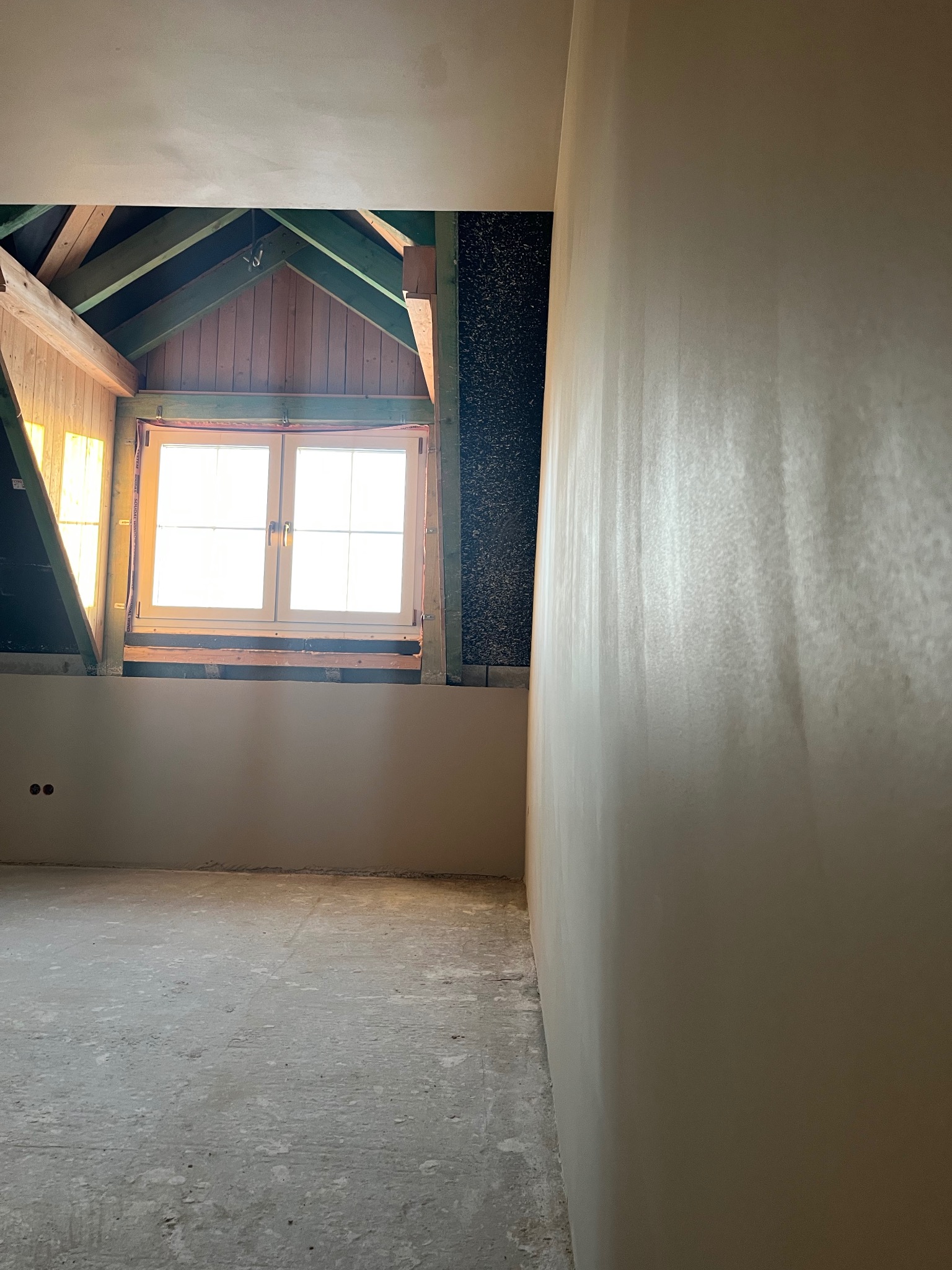 Poddasze po nałożeniu gładzi gipsowej, widoczne drewniane elementy konstrukcji dachu i okno dachowe z wpadającym światłem słonecznym.