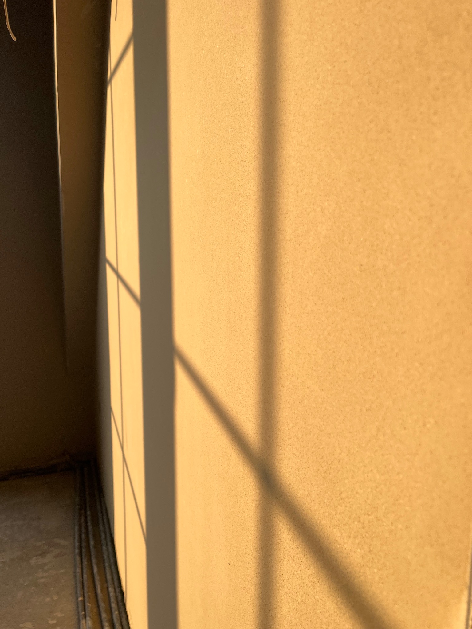 Ściana pokryta świeżym, jasnym tynkiem, oświetlona popołudniowym słońcem, rzucającym cienie okiennej ramy.