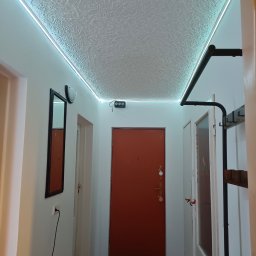 Usługi Budowlane Patryk Rożenko - Korytarz z oświetleniem LED na suficie o fakturze baranka, pomarańczowe drzwi wejściowe, lustro na ścianie i wieszak na ubrania.