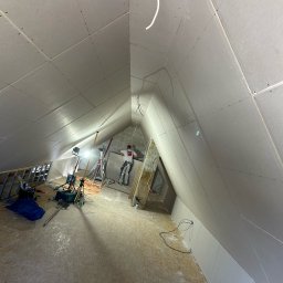 Usługi Budowlane Patryk Rożenko - Montaż płyt kartonowo-gipsowych na poddaszu, widoczne oświetlenie robocze, drabina i pracownik w tle.