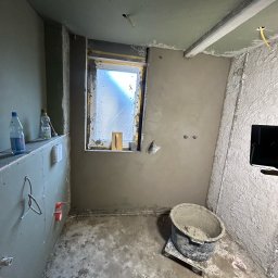 Usługi Budowlane Patryk Rożenko - Pokój w trakcie prac wykończeniowych: ściany i sufit pokryte świeżym tynkiem, widoczne butelki z wodą na półce, wiadro z zaprawą na podłodze, okno zabezpieczone folią.