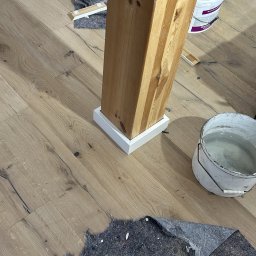 Usługi Budowlane Patryk Rożenko - Drewniany słup z białą listwą przypodłogową na nowej drewnianej podłodze, zabezpieczony materiałem ochronnym i wiadrem z farbą w tle.