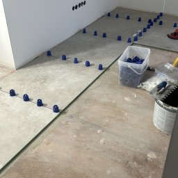 Usługi Budowlane Patryk Rożenko - Układanie płytek podłogowych z użyciem systemu poziomowania, widoczne niebieskie klipsy, poziom laserowy, pojemnik z klipsami, paca do nakładania kleju i surowa podłoga.