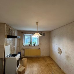 Usługi Budowlane Patryk Rożenko - Wnętrze kuchni przed remontem: widoczne stare płytki, zniszczone ściany, żółty grzejnik pod oknem, kuchenka gazowa i parkiet w jodełkę.