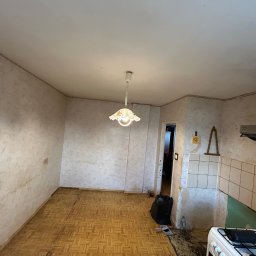 Usługi Budowlane Patryk Rożenko - Kuchnia w trakcie remontu: zerwane stare płytki, widoczne ślady po kleju na ścianach, parkiet w stanie wymagającym odnowienia, wisząca lampa z abażurem w kwiaty, śmieci w worku, stara kuchenka.