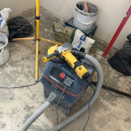 Usługi Budowlane Patryk Rożenko - Żółta szlifierka DeWalt z podłączonym odkurzaczem przemysłowym na tle worków z zaprawą, poziomicy i lampy budowlanej w trakcie prac wykończeniowych.