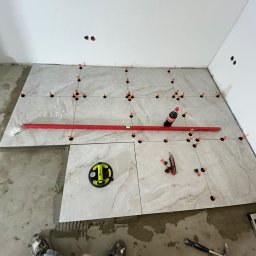 Usługi Budowlane Patryk Rożenko - Układanie płytek podłogowych w trakcie prac: poziomica, przyssawka do płytek, butelka Coca-Coli, nożyk i młotek na podłodze z częściowo ułożonymi płytkami. Widoczne spoiwa i krzyżyki dystansowe.