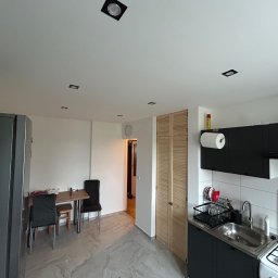 Usługi Budowlane Patryk Rożenko - Nowoczesna kuchnia z czarnymi szafkami i białymi płytkami, widoczny stół jadalny z krzesłami w tle, marmurowa podłoga i oświetlenie sufitowe.