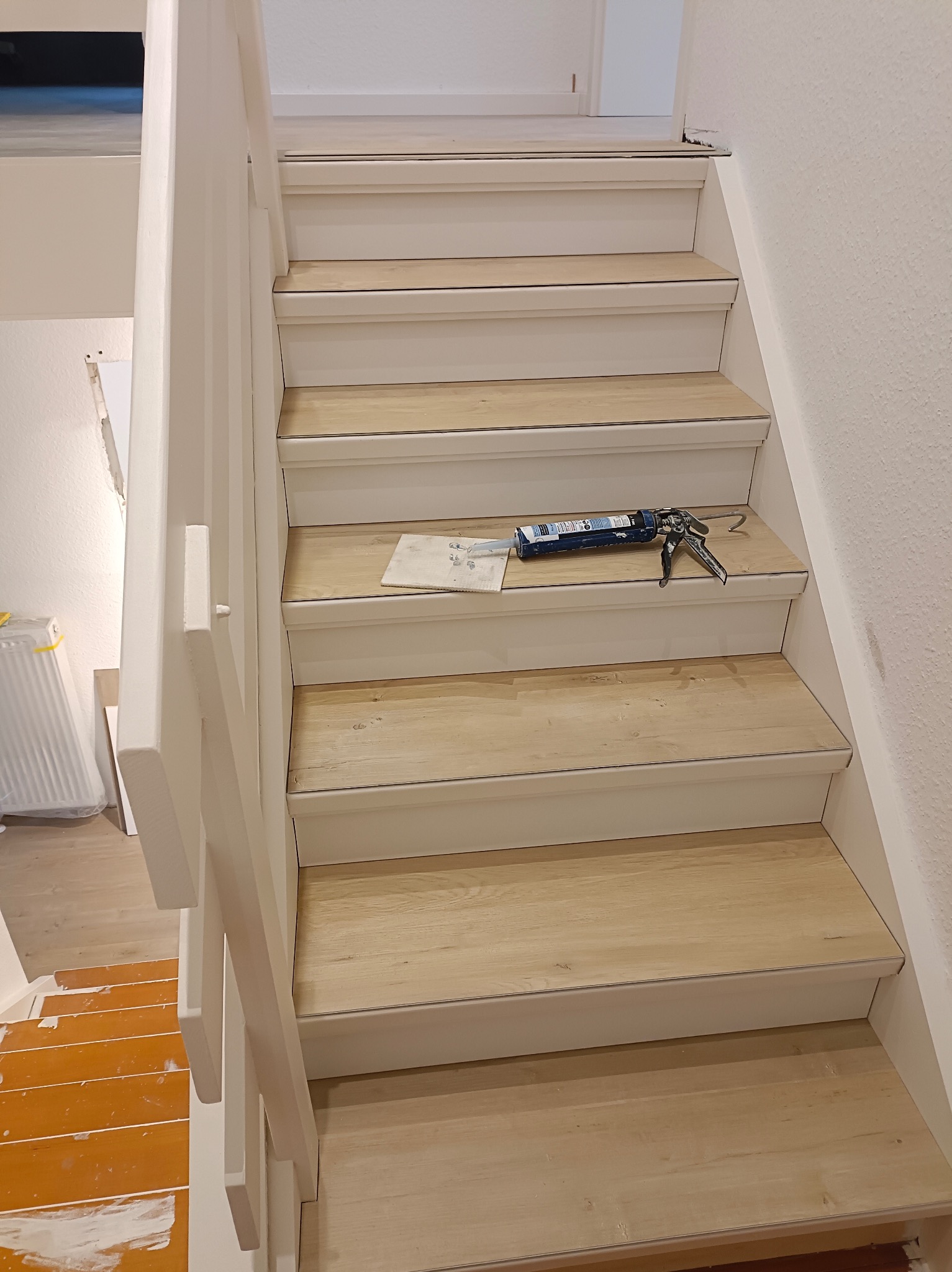 Montaż paneli na schodach z użyciem pistoletu do silikonu, widoczne narzędzia i fragment pomalowanej podłogi na dole.