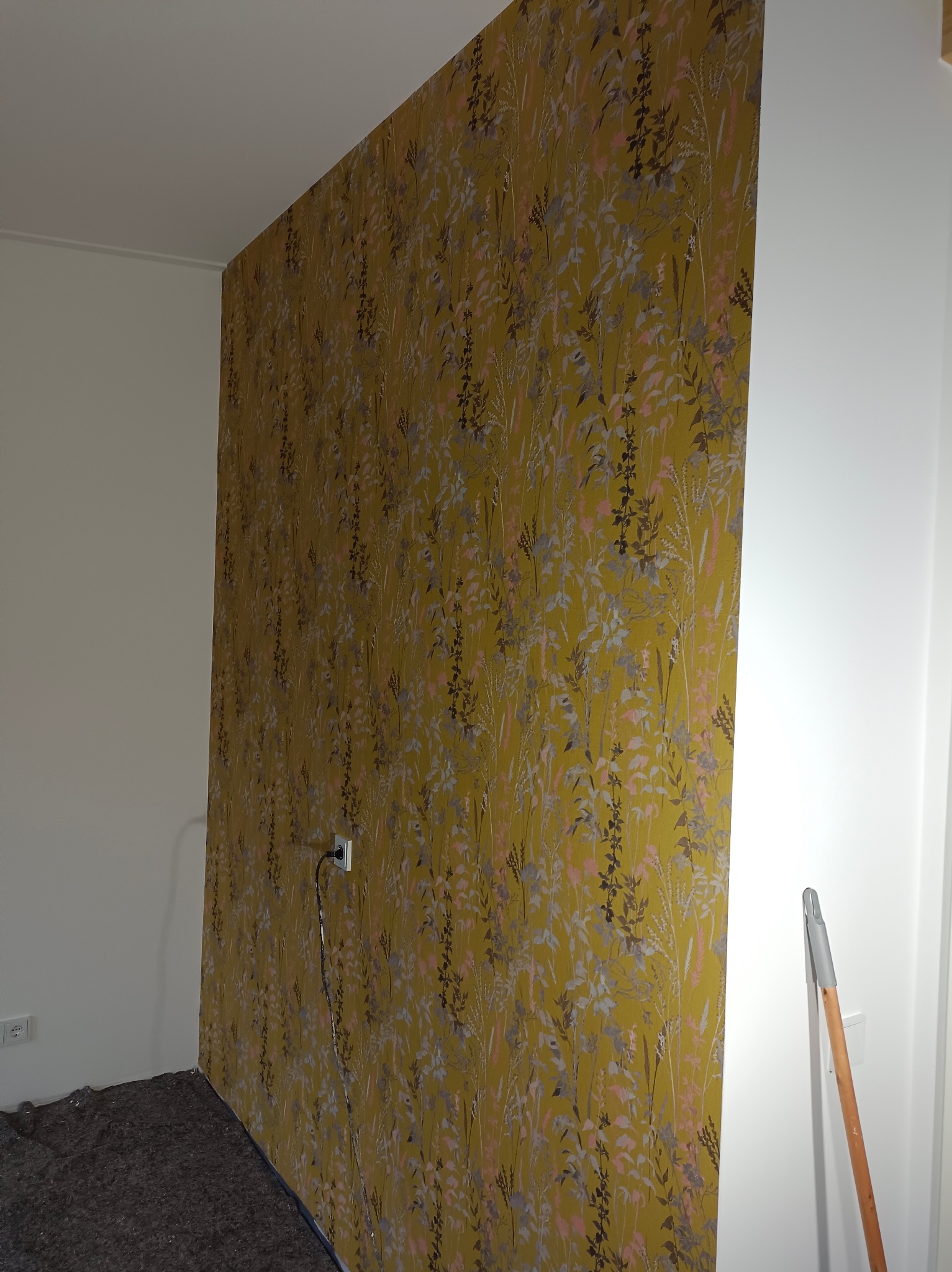 Fragment pokoju z żółtą tapetą w kwiaty, widoczne gniazdko elektryczne i narzędzie opierające się o ścianę.