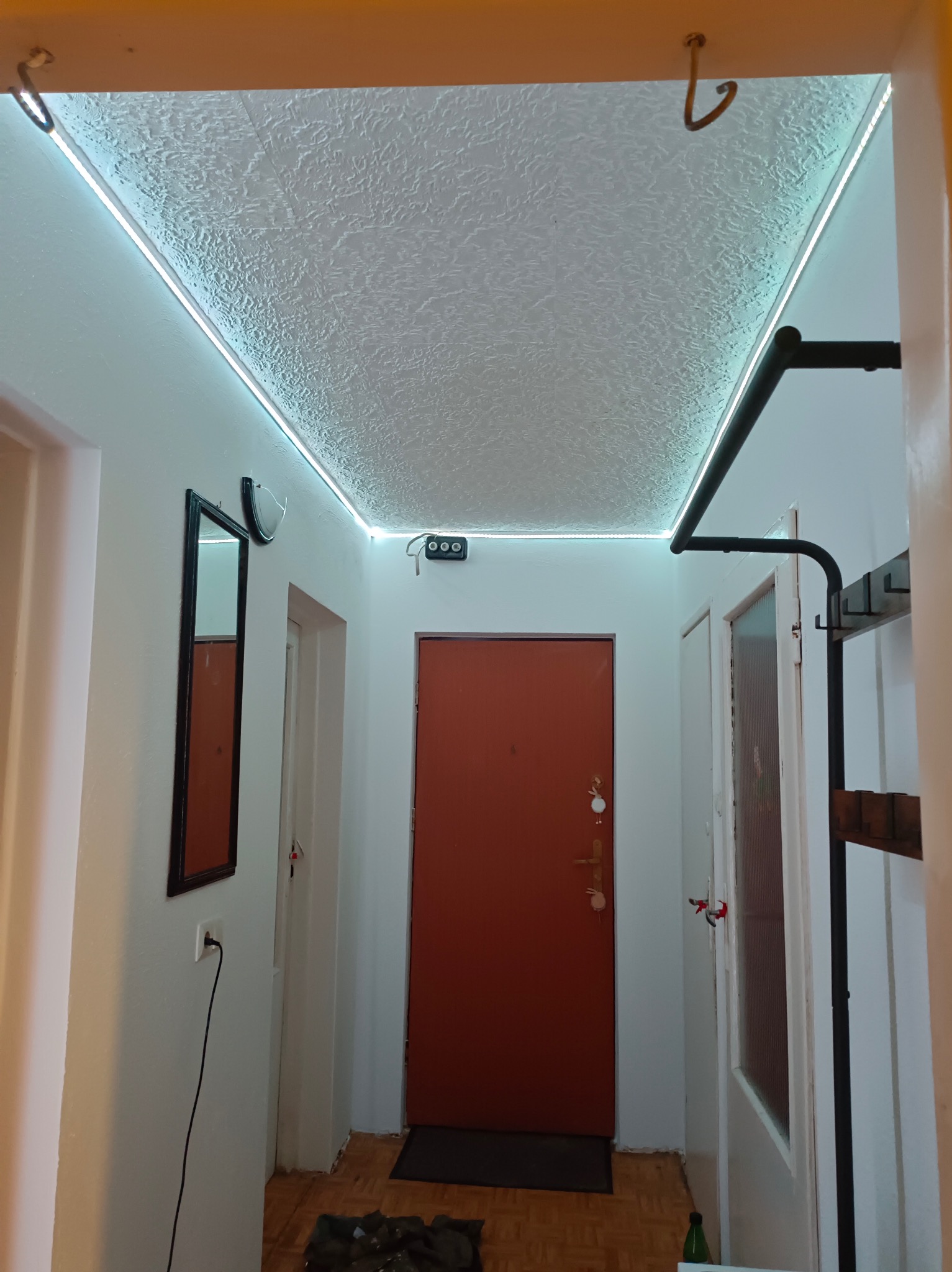 Korytarz z oświetleniem LED na suficie o fakturze baranka, pomarańczowe drzwi wejściowe, lustro na ścianie i wieszak na ubrania.