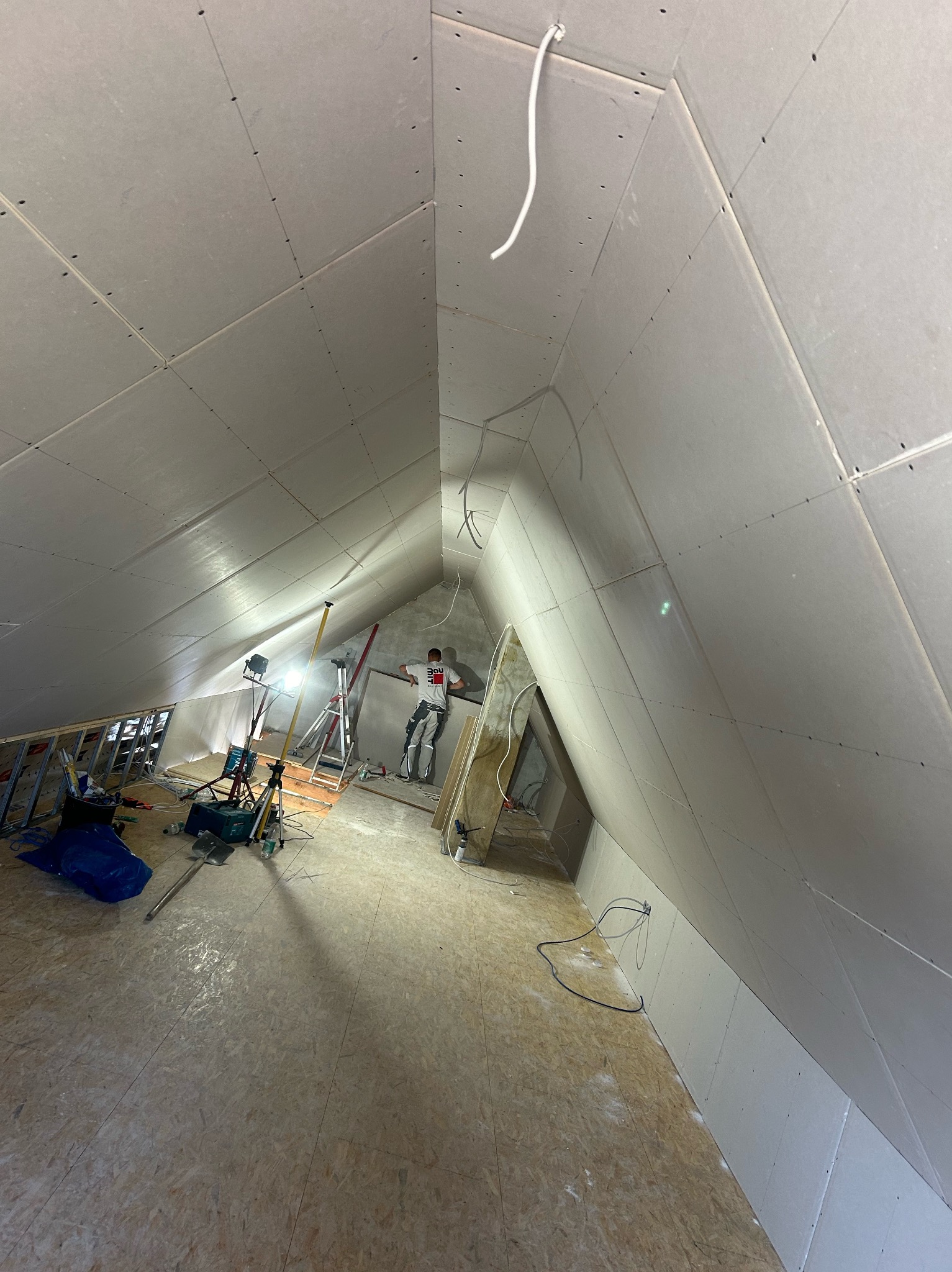 Montaż płyt kartonowo-gipsowych na poddaszu, widoczne oświetlenie robocze, drabina i pracownik w tle.